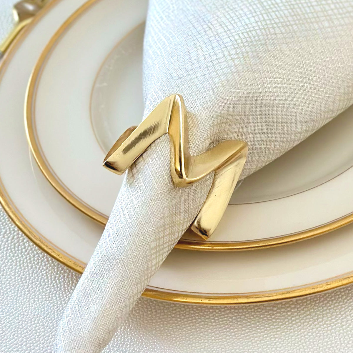 A gold-finished metal napkin ring with a bold and sculptural design made to look like a Crown. Displayed with a glistening napkin.