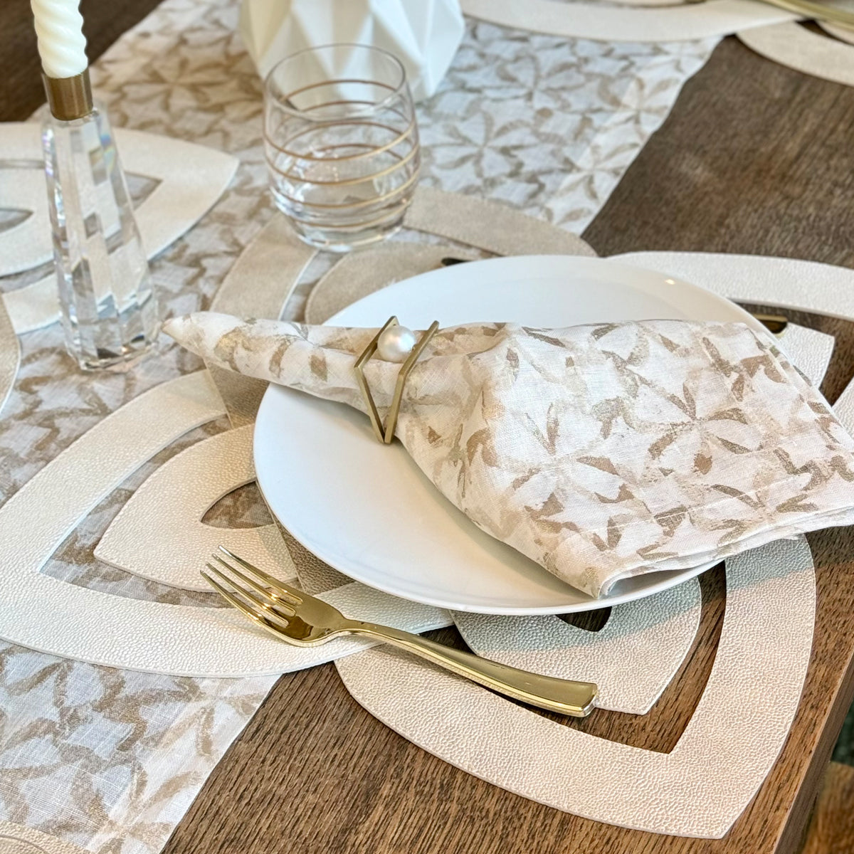Elegant table setting with white plates, gold fork, and patterned napkin on a wooden table. Focus on Napkin gold napkin ring featuring a pearl on top of it.