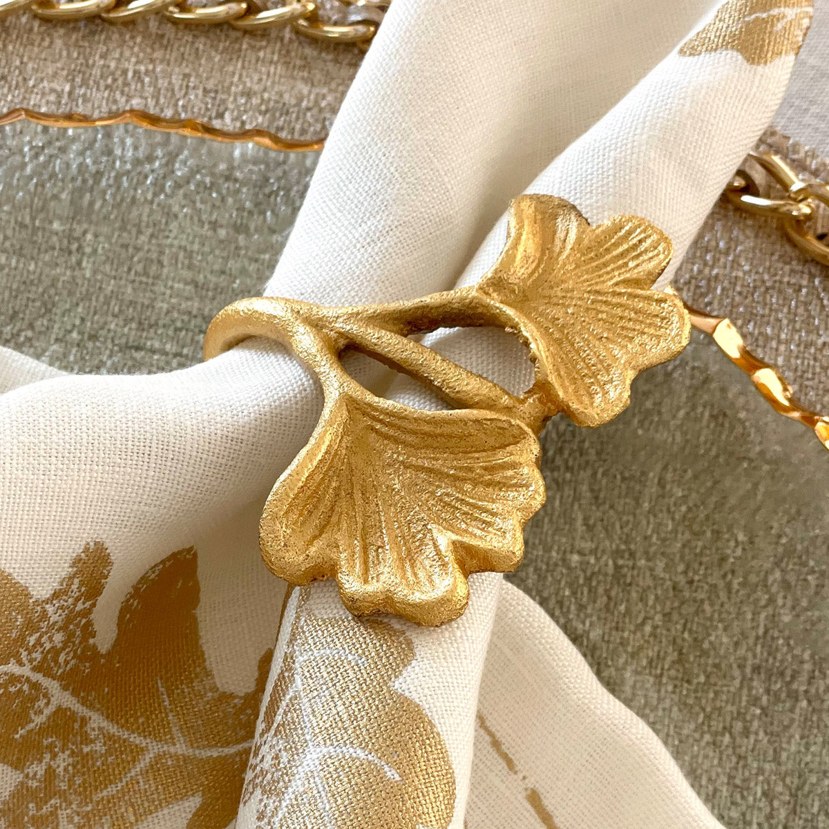 A gold-colored metal napkin ring with a ginkgo leaf design, displayed on a white napkin.