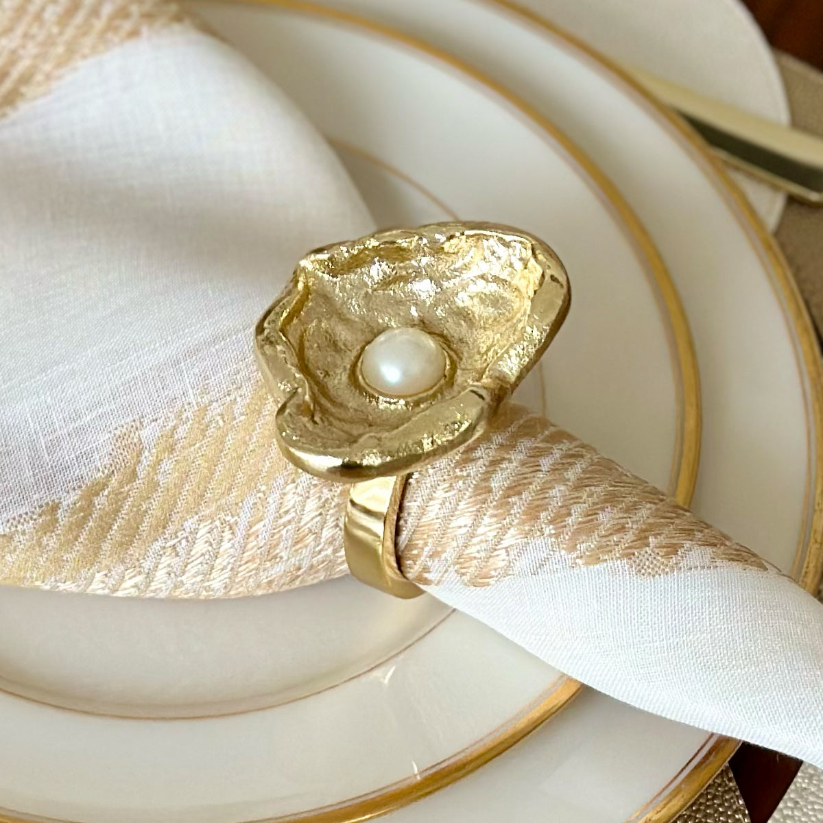 Gold napkin ring that's oyster shaped with a pearl inside on a white napkin with gold details over a gold rimmed plate