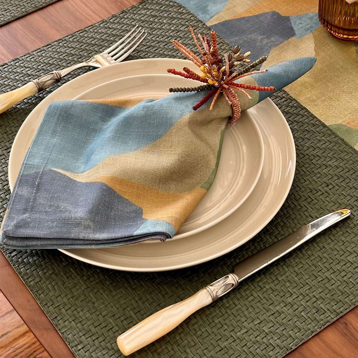 Artistic napkin with autumn inspired napkin ring on top of a oblong vinyl placemat that features a wicker look in a beautiful olive color.