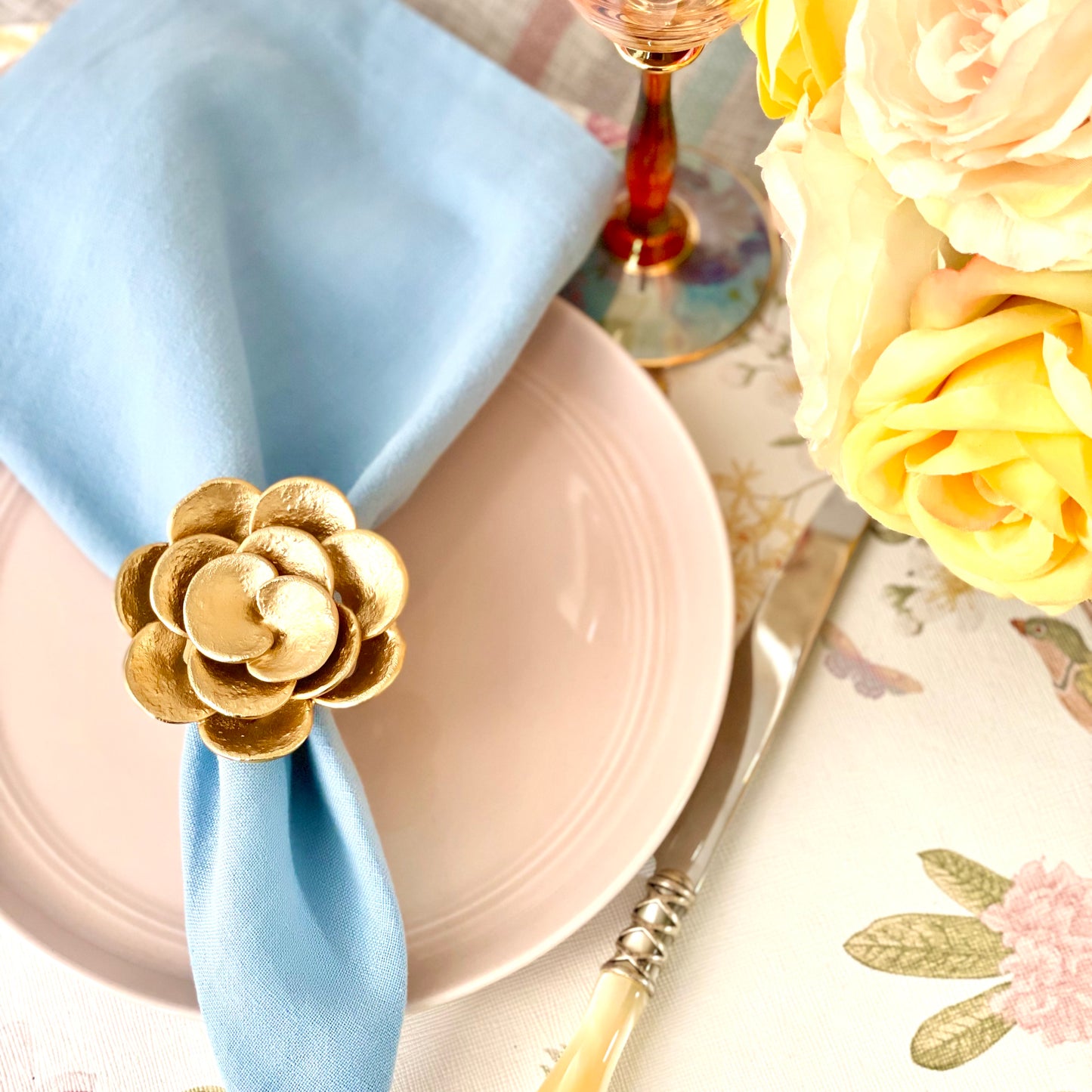 A gold metal napkin ring designed to resemble a camellia flower, displayed on a blue napkin placed on a pink plate, with a floral patterned tablecloth in the background.