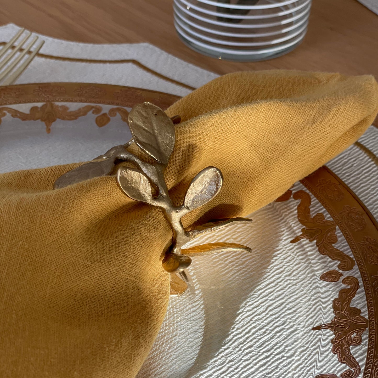 A gold-finished metal napkin ring designed to resemble laurel leaves, placed on a yellow napkin with a patterned plate in the background.
