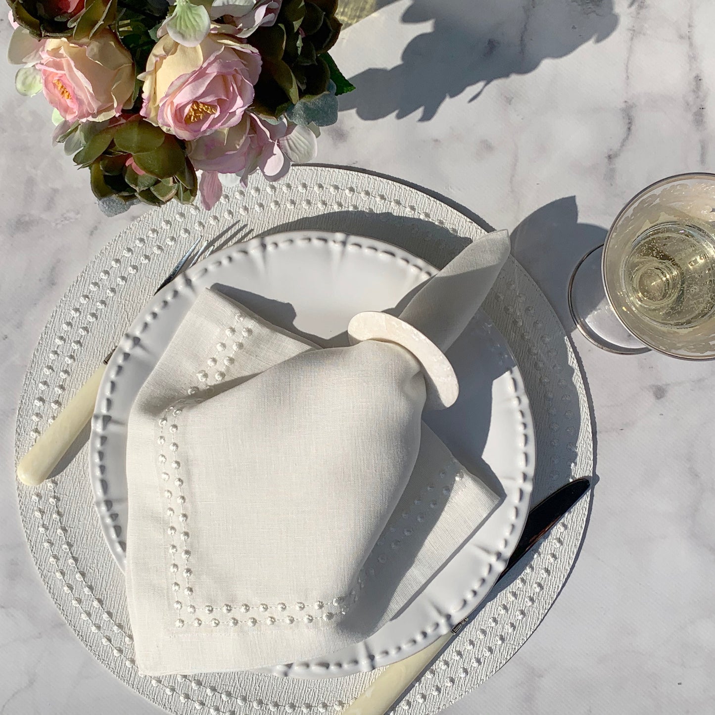 Off-white linen napkin with white 'pearl' embroidery, displayed on a plate with a flower arrangement and a glass of water in the background. Matching Pearl vinyl placemat with embroidery.
