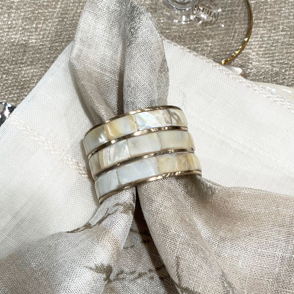 A gold-colored metal napkin ring with mother of pearl inlays, displayed on a beige linen metallic napkin.